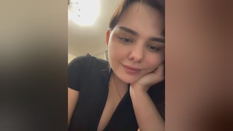 Video of a young woman with light skin and brown hair, wearing a black top, leaning on her hand, with a soft smile.