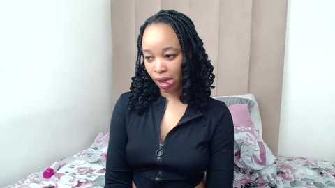 A video of a Black woman with medium skin tone, curly hair, and medium-sized breasts, wearing a black zip-up top, sitting on a bed with floral-patterned sheets, looking thoughtful.