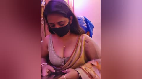 A video of a South Asian woman with medium skin tone, wearing a gray tank top revealing cleavage, a black face mask, and a yellow saree. She is focused on a smartphone, in a dimly lit, narrow room with purple and pink hues.