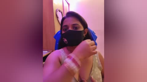 Video of a South Asian woman with dark skin and long black hair, wearing a black mask, light blue top, and a pink bracelet. She's sitting in a dimly lit room with a wooden door and a framed picture on the wall.