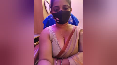 A video shows a South Asian woman with medium skin tone, wearing a black mask, blue sari, and gold jewelry. She sits in a dimly lit room with blurred objects in the background.
