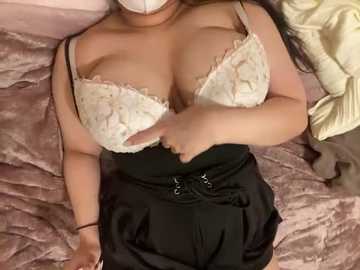 Video of a woman with medium skin tone, wearing a lacy white bra, black pleated skirt, and beige mask. She lies on a mauve quilted bedspread, partially covered by a yellow blanket.