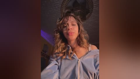 Video of a young woman with long, wavy blonde hair, wearing a light blue, off-the-shoulder shirt, leaning against a brick wall, with a dim, cozy room background.