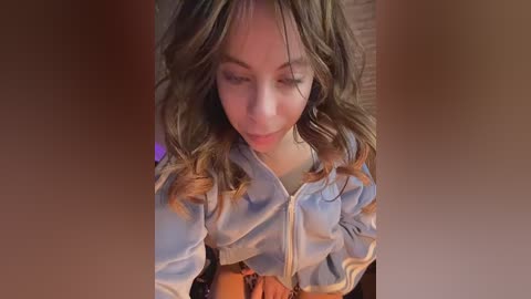 Video of a young girl with light brown, wavy hair and fair skin, wearing a light blue zip-up jacket, looking down with a neutral expression. Background is blurred, focusing attention on her.