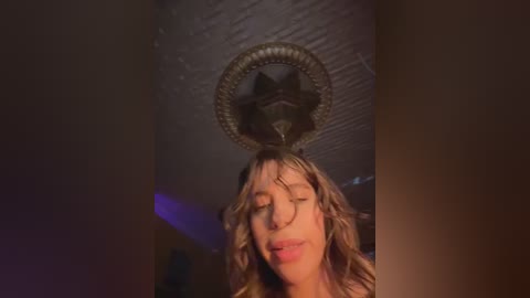 Video of a woman with long, wavy brown hair, eyes closed, wearing a black dress, standing under a chandelier with a star-shaped light fixture, in a dimly lit room with purple lighting.