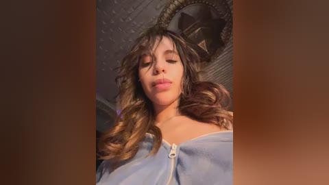 Video of a young woman with wavy brown hair, fair skin, and a light blue hoodie, standing against a dimly lit, textured background with a round mirror.