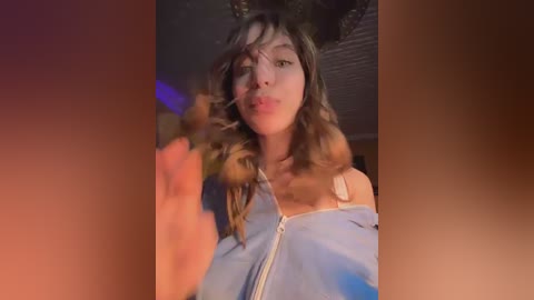 A video of a young woman with wavy, shoulder-length brown hair, wearing a blue hoodie, making a pouty face while holding her phone. The background is dimly lit with blurred purple and orange tones.
