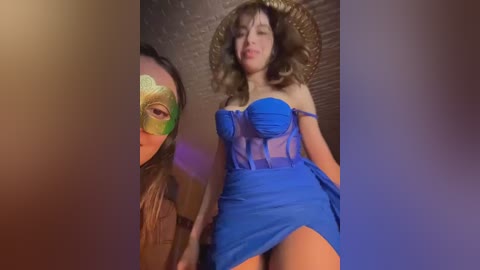 Video of two women in a dimly-lit room, one wearing a green mask, the other a blue dress with a high slit, both with wavy brown hair.