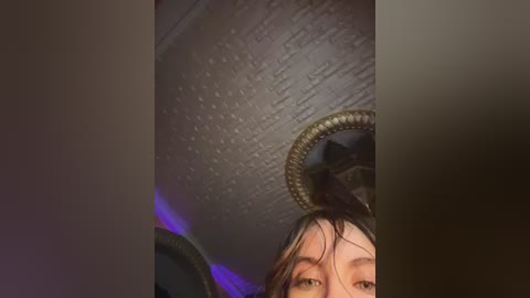 A video captures a young woman's face with wet, dark hair, partially obscured by a circular, ornate mirror. The background features a textured, metallic ceiling and purple LED lighting, suggesting an industrial or modern setting.