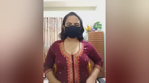Video of a woman in a maroon traditional dress with intricate embroidery, black face mask, and dark hair, sitting indoors with a wooden dresser and floral arrangement in the background.