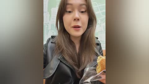 Video of an Asian woman with long brown hair, wearing a black leather jacket, holding a bagel with cream cheese, in a blurred, green subway map background.
