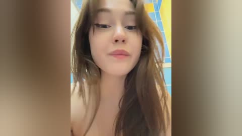 A video of a young, light-skinned woman with long, brown hair, looking downward with a neutral expression, taken in a bathroom with blue and yellow tiled walls.