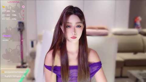 Video of a young Asian woman with long, straight brown hair, wearing a purple off-shoulder dress. She stands in a modern, minimalist living room with a beige sofa and white walls.