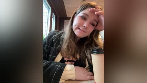 Video of a young woman with light brown hair, wearing a plaid jacket, leaning on a table with a coffee cup, indoors with wooden walls and windows.