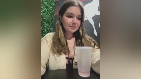 Video of a young Caucasian woman with light skin and long brown hair, wearing a cream-colored knitted cardigan over a black bra, holding a clear plastic cup. The background features a green wall and a black wall with a white silhouette.