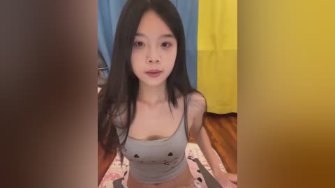 Video of an Asian woman with long black hair, wearing a gray tank top with small hearts, kneeling on a pink and white patterned mat, partially obscured by two yellow and blue curtains.