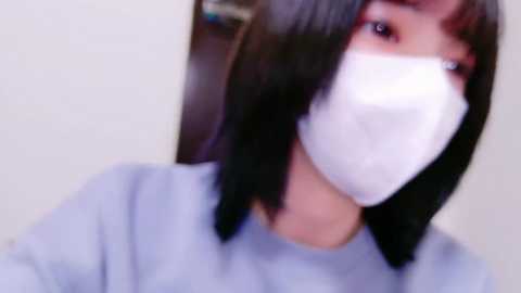 A blurry video of a young Asian woman with straight, shoulder-length black hair, wearing a white medical mask and a light blue shirt, standing against a plain, light-colored background.