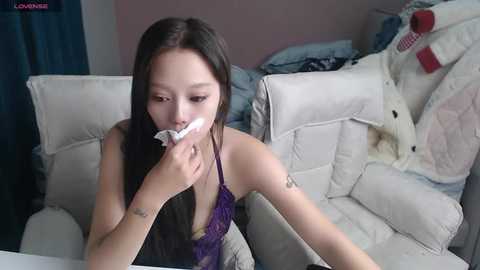 Video of an Asian woman with long black hair, wearing a purple sleeveless dress, blowing her nose on a tissue, seated on a white leather couch in a messy room with clothes and a stuffed animal.