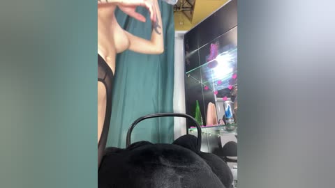 A video of a topless woman with fair skin and a slender physique, wearing black lingerie, standing in a dimly lit room with a teal curtain, a glass aquarium, and various sex toys on display.