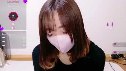A video of an East Asian woman with straight, shoulder-length brown hair, wearing a light pink face mask and black top, standing indoors against a white wall with pink silhouettes and a video game UI in the background.