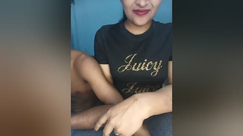 Video of a smiling Asian woman with medium skin tone, wearing a black t-shirt with \"Juniors\" in gold letters, sitting on a blue chair with a leg on a grey sofa.