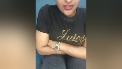 Video of a young woman with light skin and a slim build, wearing a black T-shirt with \"Lucky\" in glittery gold letters, gray leggings, and a silver wristwatch. She sits cross-legged on a blue couch, mouth slightly open, showing teeth.