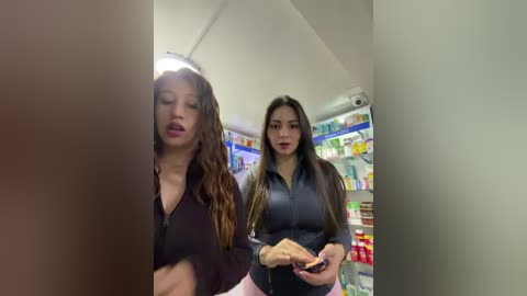 A video of two young women, one with long brown hair, wearing a black top, and another with long straight hair, in a brightly lit convenience store, holding a phone.