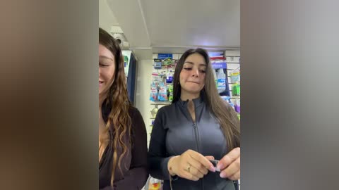 Video of two women in a pharmacy; one with wavy hair, wearing a black top, the other with straight hair, in a black zip-up. Shelves filled with various products in the background.