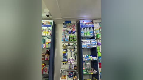 Video of a brightly lit pharmacy aisle, showcasing neatly organized shelves filled with various medications, vitamins, and first aid supplies. The background features a white wall and ceiling.