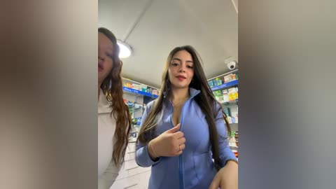 Video of a young woman with long brown hair and fair skin, dressed in a light blue blouse, standing in a pharmacy, gesturing with her right hand.