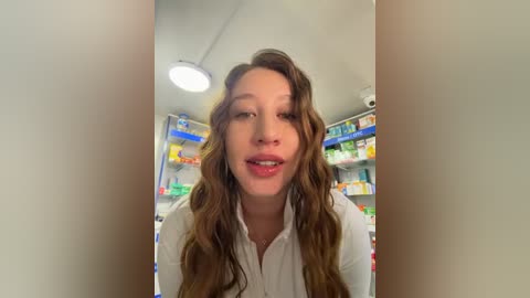Video of a Caucasian woman with long, wavy brown hair and light skin, wearing a white blouse, smiling in a brightly lit pharmacy with colorful medicine shelves in the background.