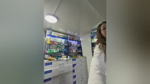 A video of a woman with long, wavy hair partially visible, standing in a brightly lit, modern pharmacy. The shelves are stocked with various medications and products.