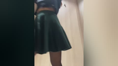 Video of a person from the back, wearing a dark green, short, pleated skirt and a cropped top, standing in a narrow hallway with cream-colored walls.