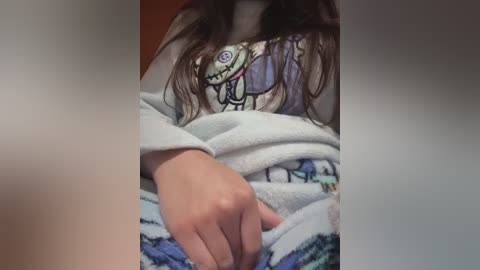 Video of a person's hand resting on a white blanket with a colorful, cartoonish elephant pattern, partially covered by a gray sweatshirt. The background is blurred, focusing attention on the hand and fabric textures.