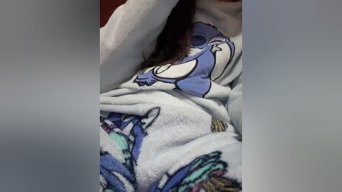 A video of a person in a white plushy sweater featuring a cartoonish blue bird design. The person's face is partially obscured, and they are seen from the waist up, with long dark hair partially visible. The background is blurry and nondescript.