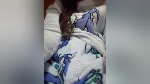 Video of a person in a blue and white unicorn-themed pajama top, showcasing a cartoon unicorn design. The individual's long brown hair is partially visible. The background is blurred, with a soft gradient of gray tones.