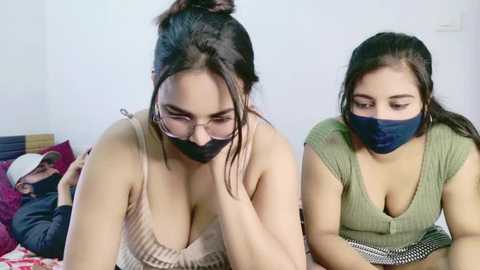 A video of two women with medium skin tones, wearing face masks, sitting on a bed. One has black hair in a bun, wearing glasses and a beige top, looking tired. The other has dark hair, a green top, and a mask, looking contemplative.