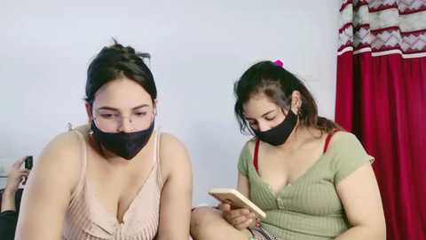 Video of two women in casual attire, wearing black face masks, sitting in a room with white walls and red curtains, focused on their phones.
