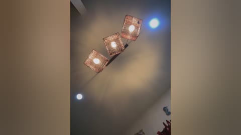 Video of a modern room ceiling with three square, geometric, red-hued pendant lights emitting warm white light. The ceiling is beige, with two additional circular lights visible, creating a cozy ambiance.