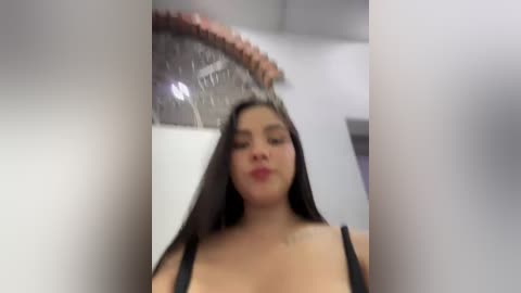 A video of a young woman with long black hair, wearing a black tank top, standing in a bathroom, looking down. The background features a showerhead and white walls.