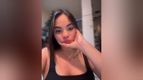 Video of a Latina woman with medium skin tone, straight dark hair, and prominent facial features, wearing a black tank top, touching her face. Background shows a blurred indoor setting with a Christmas tree and other decorations.