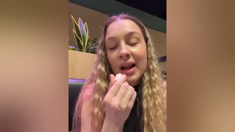 Video of a young woman with wavy blonde hair, wearing a black top, playfully sticking her tongue out while holding a finger to her mouth, set in a modern indoor environment with a potted plant.