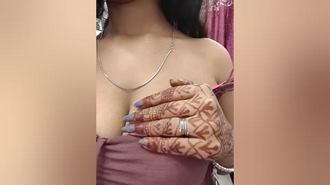 Video of a South Asian woman with light brown skin, wearing a pink off-shoulder top, showing henna designs on her hand, holding her breast, and a silver necklace.