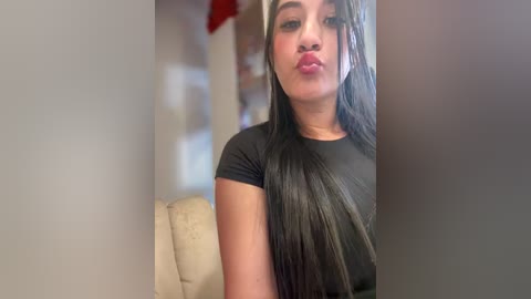 Video of a young woman with medium skin tone, long black hair, and full lips, wearing a black top. She is indoors, partially obscured by a beige couch.