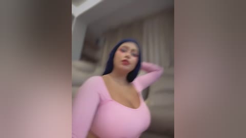 A blurred video of a Latina woman with blue hair and fair skin, wearing a tight pink long-sleeve top, posing indoors.