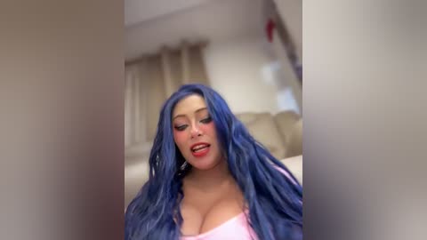 Video of a woman with long, wavy, blue hair, wearing a pink top, smiling in a blurred indoor setting.