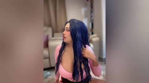 Video of a young woman with long, wavy blue hair, wearing a pink off-shoulder top, sitting on a beige couch in a modern living room.
