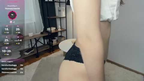 Video of a light-skinned woman wearing black lace panties, standing in a modern, minimalist room with a white shelf, white curtains, and wooden floor.