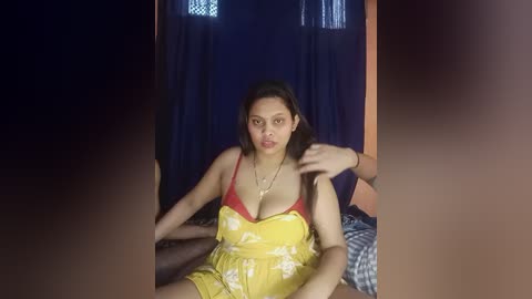 A video of an Asian woman with medium-length black hair, wearing a yellow floral dress with a red bralette, sitting on a bed in a dimly lit room with dark curtains.