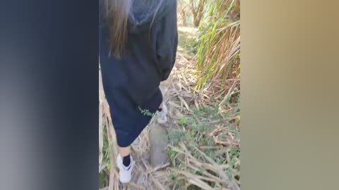 Video of a person with long hair wearing dark pants and white sneakers, standing in a sunlit, dry, grassy area. The background features tall, golden-brown grass and scattered twigs.